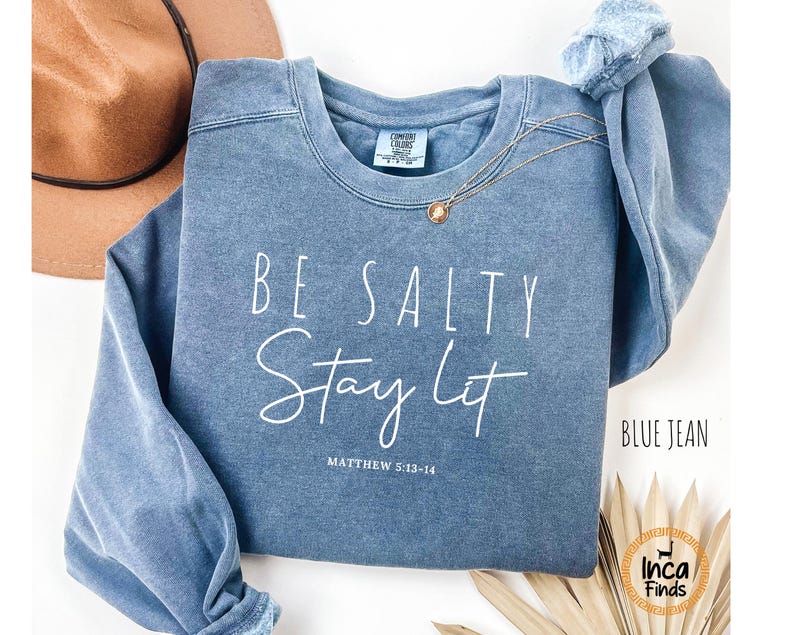 May include: A blue jean-colored sweatshirt with the words "BE SALTY Stay Lit" in white script. The text "MATTHEW 5:13-14" is below the main text. A gold necklace and a brown hat are also visible.