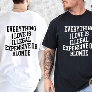 Pu&ograve; includere: Due magliette, una bianca e una nera, ciascuna con la frase "EVERYTHING I LOVE IS ILLEGAL EXPENSIVE OR BLONDE" stampata in grassetto bianco. La maglietta bianca &egrave; a sinistra, e quella nera &egrave; a destra.
