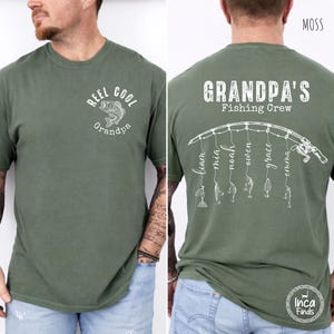 May include: A moss green short-sleeve t-shirt featuring white graphics. The front displays a fish and the text "REEL COOL Grandpa". The back shows a fishing rod with lines hanging down, each ending in a lure and a name, with the text "GRANDPA'S Fishing Crew".