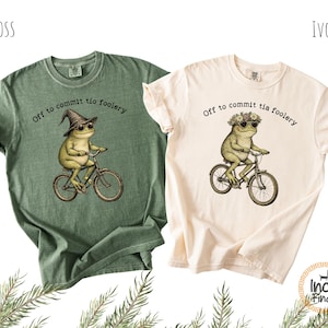 Puede incluir: Dos camisetas, una verde musgo y otra marfil, con una ilustración caprichosa de una rana en bicicleta. La rana lleva gafas de sol y un sombrero o corona de flores, con el texto "Off to commit tio foolery". Las camisetas son informales.