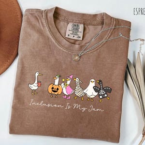 Peut inclure: T-shirt couleur expresso avec un col rond et le texte "Inclusion Is My Jam". Le t-shirt présente une ligne de canards de dessins animés dans divers costumes, dont une citrouille, une sorcière et une momie. Un collier en argent avec un pendentif est drapé sur le t-shirt.
