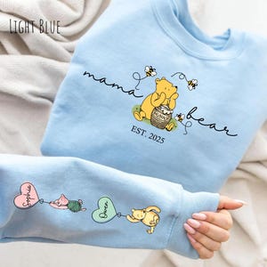 Könnte beinhalten: Hellblauer Pullover mit dem Text "mama bear est. 2025" und einem Winnie-Puuh-Cartoon, der einen Honigtopf hält. Der Ärmel hat ein rosa Herz mit dem Text "Sophia" und ein grünes Herz mit dem Text "Gwen" mit Winnie-Puuh-Cartoon-Charakteren.