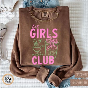 May include: Espresso-colored long-sleeve tee with the text "hot GIRLS matcha CLUB" in pink and green. The graphic includes a matcha bowl, stars, and a matcha drink with ice cubes. A gold chain necklace is visible.