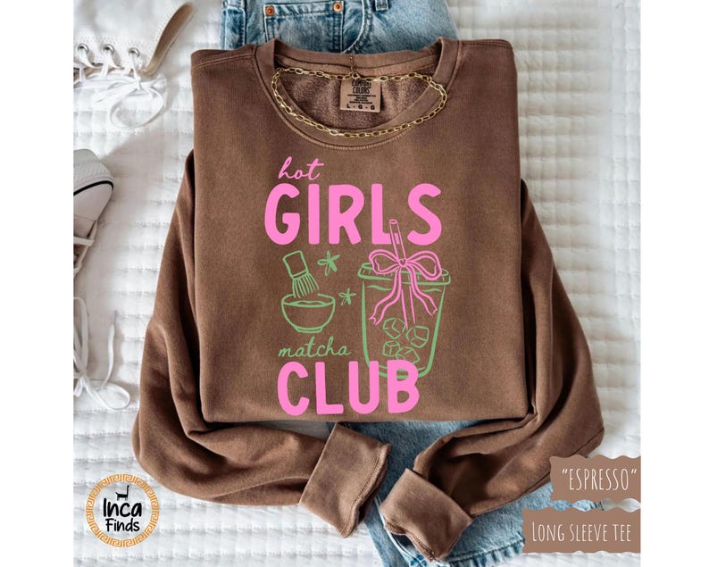 May include: Espresso-colored long-sleeve tee with the text "hot GIRLS matcha CLUB" in pink and green. The graphic includes a matcha bowl, stars, and a matcha drink with ice cubes. A gold chain necklace is visible.
