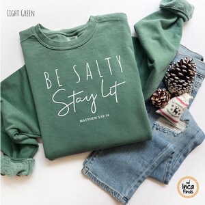 May include: A light green sweatshirt with the text "BE SALTY Stay Lit" in white script. The sweatshirt is paired with blue jeans, pine cones, and a small decorative bag. The text "Matthew 5:13-14" is below the main text.