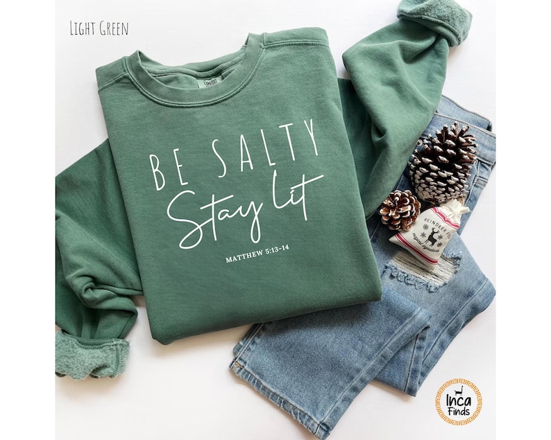 May include: A light green sweatshirt with the text "BE SALTY Stay Lit" in white script. The sweatshirt is paired with blue jeans, pine cones, and a small decorative bag. The text "Matthew 5:13-14" is below the main text.