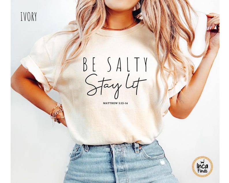 May include: Ivory-colored t-shirt with the text "BE SALTY Stay Lit" in black script. The shirt also includes the Bible verse Matthew 5:13-14. The shirt is paired with blue denim shorts.