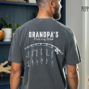 May include: A charcoal gray short-sleeve t-shirt featuring a white graphic on the back. The design displays a fishing rod with six hanging lines, each ending in a fishing lure and a name: Liam, Mia, Noah, Owen, Grace, and Emma. Above the rod, the text reads "GRANDPA'S Fishing Crew" in a distressed white font.