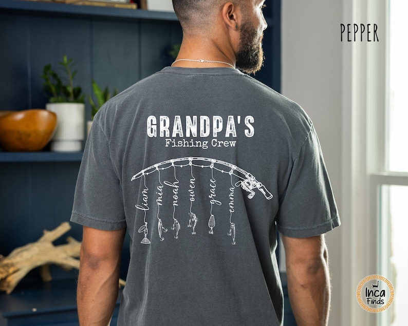 May include: A charcoal gray short-sleeve t-shirt featuring a white graphic on the back. The design displays a fishing rod with six hanging lines, each ending in a fishing lure and a name: Liam, Mia, Noah, Owen, Grace, and Emma. Above the rod, the text reads "GRANDPA'S Fishing Crew" in a distressed white font.