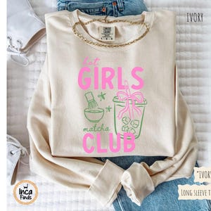 May include: Ivory long-sleeve tee with the text "Hot Girls Matcha Club" in pink and green. The design includes matcha tools and a matcha drink with ice cubes. A gold chain necklace is visible.