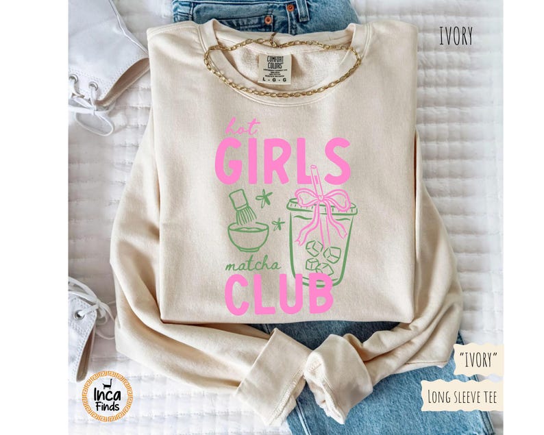 May include: Ivory long-sleeve tee with the text "Hot Girls Matcha Club" in pink and green. The design includes matcha tools and a matcha drink with ice cubes. A gold chain necklace is visible.