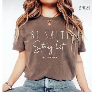 May include: Espresso-colored t-shirt with the text "BE SALTY Stay Lit" in white script. The text "Matthew 5:13-14" is below the script. The shirt is a short-sleeved, crew neck style.