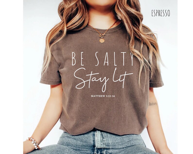 May include: Espresso-colored t-shirt with the text "BE SALTY Stay Lit" in white script. The text "Matthew 5:13-14" is below the script. The shirt is a short-sleeved, crew neck style.
