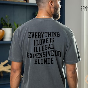 May include: Pepper-colored t-shirt with the text "EVERYTHING I LOVE IS ILLEGAL EXPENSIVE OR BLONDE" in black block letters. The shirt is a casual style, likely made of soft cotton, and has a vintage, worn look.
