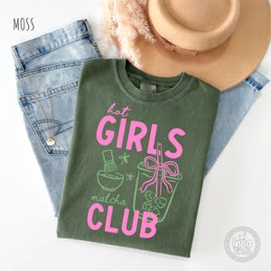 May include: An olive green t-shirt with the text "hot GIRLS matcha CLUB" in pink. The shirt features a matcha bowl and a matcha drink graphic. The shirt is laid on top of a pair of blue jeans.