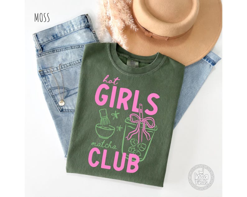May include: An olive green t-shirt with the text "hot GIRLS matcha CLUB" in pink. The shirt features a matcha bowl and a matcha drink graphic. The shirt is laid on top of a pair of blue jeans.