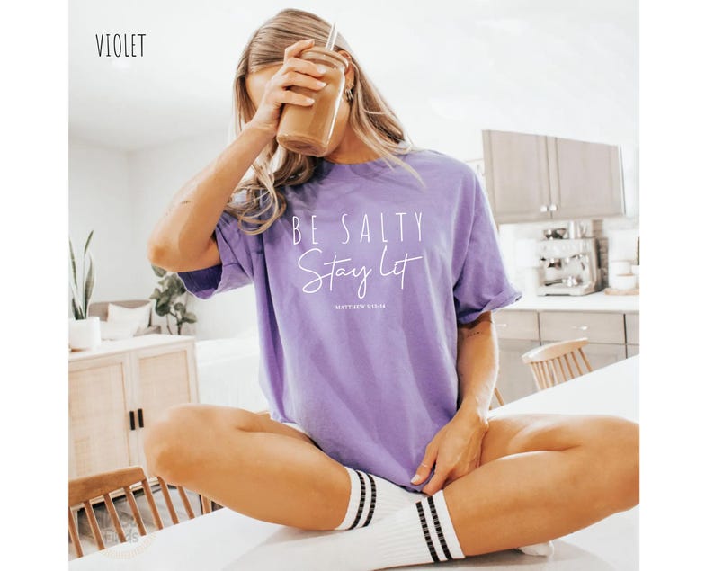 May include: A violet-colored t-shirt with the text "BE SALTY Stay Lot" and "Matthew 5:13-16" printed in white. The shirt is worn with white socks with black stripes. The word "VIOLET" is in the upper left corner.