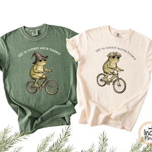 Puede incluir: Dos camisetas, una verde y otra crema, cada una con una rana en bicicleta. La camiseta verde tiene una rana con un sombrero de bruja, y la camiseta crema tiene una rana con una corona de flores. Ambas camisetas tienen el texto "Off to commit uncle/auntie foolery."