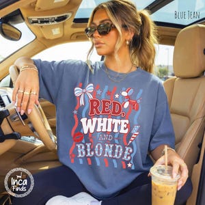 May include: A blue jean-coloured t-shirt with the words "RED, WHITE, and BLONDE" in a patriotic design. The shirt features red, white, and blue graphics, including a bow, stars, and a lipstick kiss. The shirt is worn in a car.