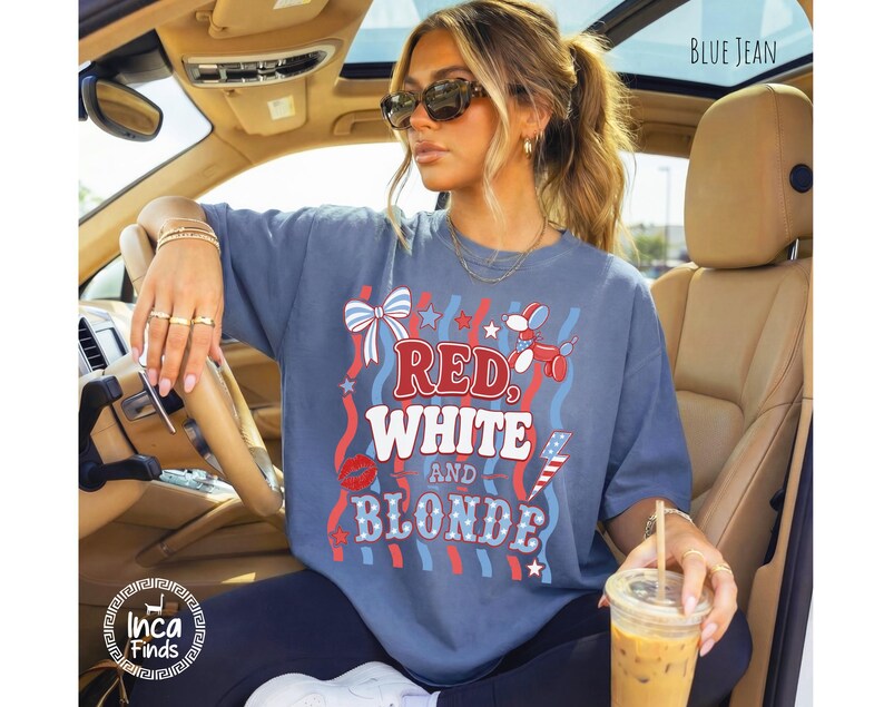 May include: A blue jean-coloured t-shirt with the words "RED, WHITE, and BLONDE" in a patriotic design. The shirt features red, white, and blue graphics, including a bow, stars, and a lipstick kiss. The shirt is worn in a car.
