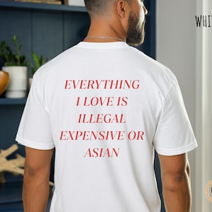 Op de afbeelding: Wit T-shirt met rode tekst: "EVERYTHING I LOVE IS ILLEGAL EXPENSIVE OR ASIAN." Het woord "WHITE" is in het zwart rechtsboven gedrukt. Het shirt heeft een casual stijl.