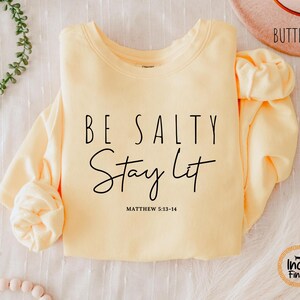 May include: A light yellow sweatshirt with the text "BE SALTY Stay Lit" in black script. The sweatshirt also includes the Bible verse Matthew 5:13-14. The sleeves are folded, and the word "Butter" is visible on a hat in the background.