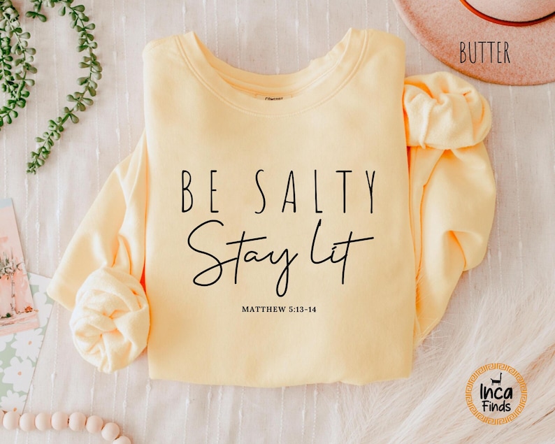 May include: A light yellow sweatshirt with the text "BE SALTY Stay Lit" in black script. The sweatshirt also includes the Bible verse Matthew 5:13-14. The sleeves are folded, and the word "Butter" is visible on a hat in the background.