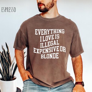 Pu&ograve; includere: T-shirt color espresso con la scritta bianca "EVERYTHING I LOVE IS ILLEGAL EXPENSIVE OR BLONDE". La maglietta ha una vestibilit&agrave; ampia e viene indossata.