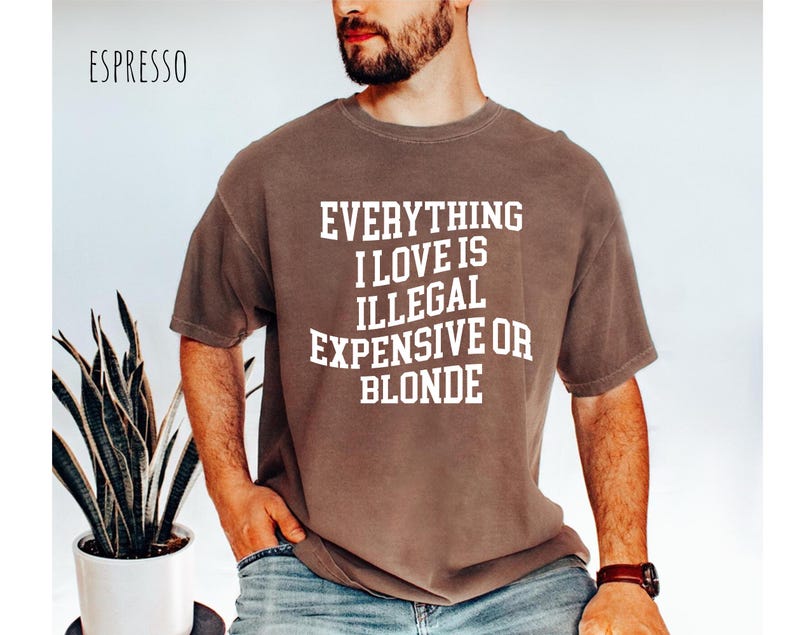 Pu&ograve; includere: T-shirt color espresso con la scritta bianca "EVERYTHING I LOVE IS ILLEGAL EXPENSIVE OR BLONDE". La maglietta ha una vestibilit&agrave; ampia e viene indossata.