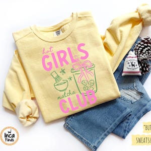May include: A light yellow sweatshirt with the text "hot GIRLS matcha CLUB" in pink and green. The sweatshirt is paired with blue jeans, a pinecone, and a small bag.