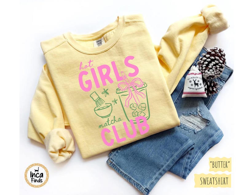 May include: A light yellow sweatshirt with the text "hot GIRLS matcha CLUB" in pink and green. The sweatshirt is paired with blue jeans, a pinecone, and a small bag.