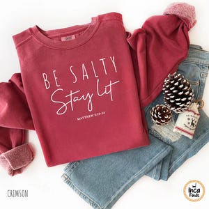 May include: Crimson-colored sweatshirt with the text "BE SALTY Stay Lit" and the Bible verse Matthew 5:13-14. The sweatshirt is paired with blue jeans, pine cones, and a small gift bag.
