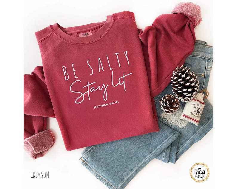 May include: Crimson-colored sweatshirt with the text "BE SALTY Stay Lit" and the Bible verse Matthew 5:13-14. The sweatshirt is paired with blue jeans, pine cones, and a small gift bag.