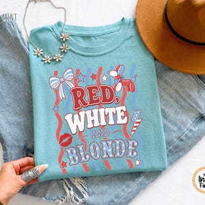 May include: A turquoise t-shirt featuring the text "RED, WHITE AND BLONDE" in red and white, with patriotic designs. A brown hat and blue jeans are also visible in the image.