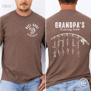 May include: An espresso brown t-shirt featuring white graphics. The front displays a fish and the text "REEL COOL Grandpa". The back shows a fishing rod graphic with lines hanging down, each ending in a lure and the names Liam, Mia, Noah, Owen, Grace, and Emma, with the text "GRANDPA'S Fishing Crew" above.