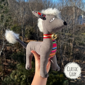May include: Gray plush toy dog with white fur on its head and tail. The dog is wearing a colorful striped collar with a gold bell.