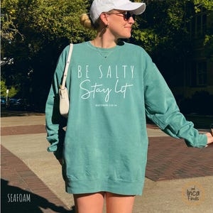May include: Seafoam green sweatshirt with the text "BE SALTY Stay Lit" in white script. The sweatshirt has a crew neck and long sleeves. The text "Matthew 5:13-16" is below the main text.