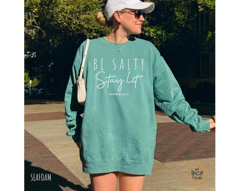 May include: Seafoam green sweatshirt with the text "BE SALTY Stay Lit" in white script. The sweatshirt has a crew neck and long sleeves. The text "Matthew 5:13-16" is below the main text.