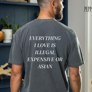 Op de afbeelding: Donkergrijs t-shirt met de witte tekst "EVERYTHING I LOVE IS ILLEGAL EXPENSIVE OR ASIAN". Het shirt heeft een relaxte pasvorm en korte mouwen. Het woord "Pepper" staat rechtsboven.