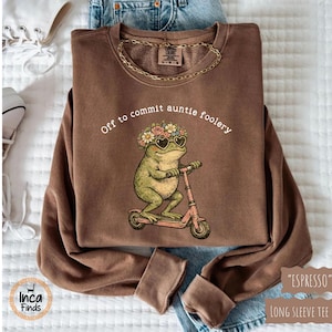 May include: Espresso-colored long-sleeve tee featuring a frog wearing sunglasses and a floral crown, riding a pink scooter. The text "Off to commit auntie foolery" is printed above the frog. A gold chain necklace is visible.