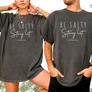 May include: Two dark gray t-shirts with the text "BE SALTY Stay Lit" and "MATTHEW 5:13-14" printed in white. The shirts have short sleeves and a relaxed fit. The word "PEPPER" is printed in the top right corner.