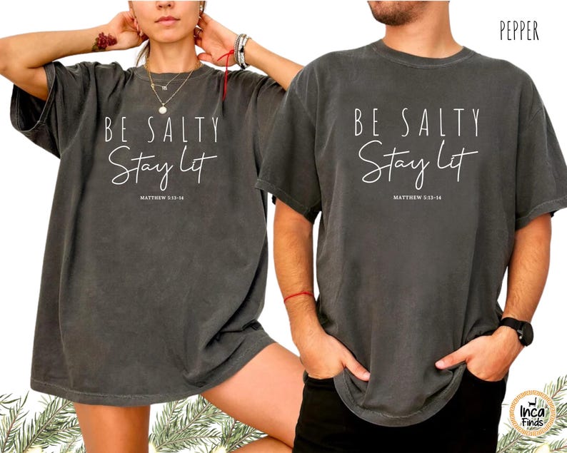 May include: Two dark gray t-shirts with the text "BE SALTY Stay Lit" and "MATTHEW 5:13-14" printed in white. The shirts have short sleeves and a relaxed fit. The word "PEPPER" is printed in the top right corner.