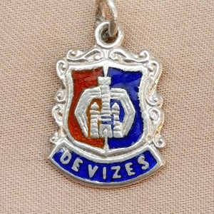 Puede incluir: Colgante de plata con diseño de escudo. El escudo está dividido en secciones rojas y azules, con un edificio estilizado. La palabra "DEVIZES" está escrita en azul sobre una pancarta curva debajo.