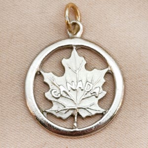 Op de afbeelding: Zilverkleurige hanger met een esdoornbladontwerp en het woord "CANADA" gegraveerd. De hanger is cirkelvormig met een kleine lus aan de bovenkant voor een ketting. Het esdoornblad is de centrale focus, met gedetailleerde nerven.