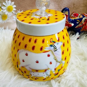 May include: A yellow ceramic jar with a white lid and a clear glass knob. The jar is decorated with a white cow and calf design, and red paisley patterns. The lid also features a cow design.