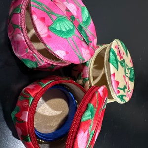 May include: Three round fabric jewellery boxes with floral patterns. The boxes are pink, beige, and red with green floral designs. The boxes have zips and are open to reveal the interior.