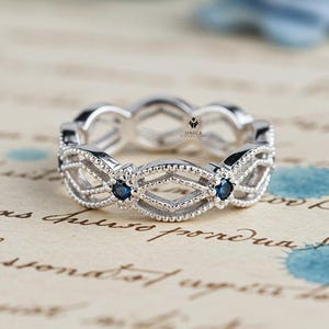 May include: A silver ring with an intricate openwork design. The ring features diamond-shaped and beaded elements, with small blue gemstones set at intervals. The ring is displayed on a vintage paper background.