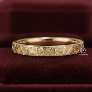 Puede incluir: Un anillo dorado con un diseño floral. El anillo se muestra sobre un fondo de terciopelo rojo oscuro. La banda está adornada con un patrón repetido de flores y hojas. La marca "onicajewels" es visible en el anillo.