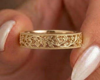 May include: A gold band ring featuring an intricate, continuous relief pattern of small leaves and flowers. The design is bordered by fine milgrain detailing along both the top and bottom edges, creating a textured, metallic finish.