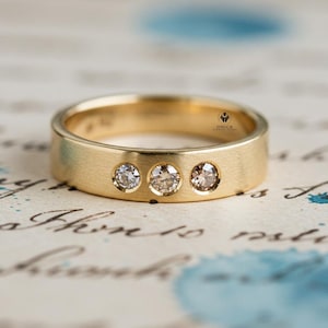May include: A gold ring featuring three clear gemstones. The ring has a brushed finish and is set against a backdrop of vintage paper with blue ink stains and handwritten script. The ring is a piece of jewellery.